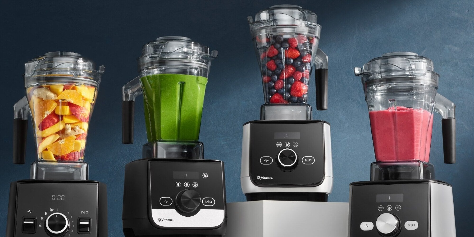Vier Vitamix Standmixer in einer Reihe, gefüllt mit verschiedenen bunten Smoothies und Früchten.