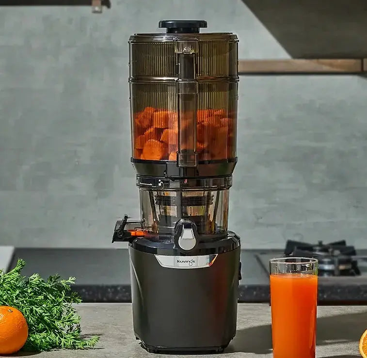 Kuvings AUTO10S Plus Entsafter gefüllt mit Karotten, daneben ein Glas frischer Karottensaft.