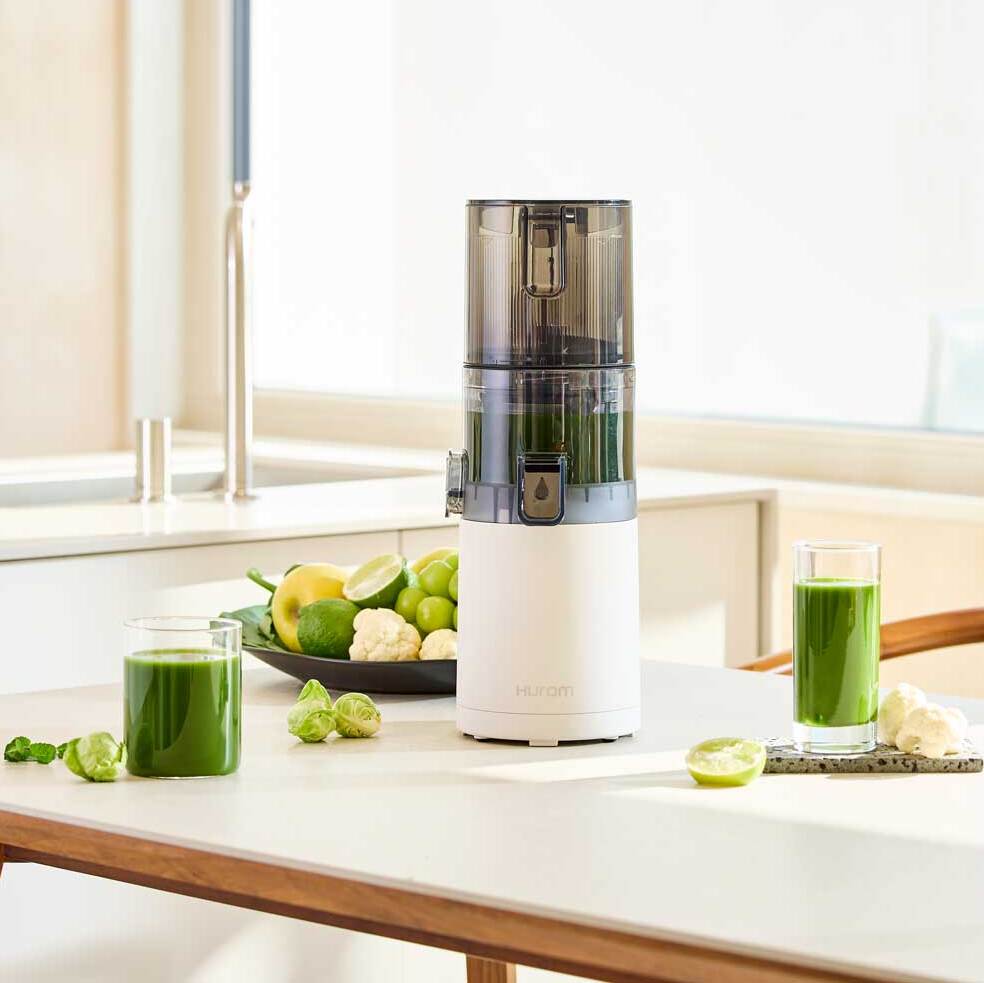 Hurom E30-ST Entsafter in moderner Küche, umgeben von grünem Gemüse, Obst und zwei Gläsern Saft.