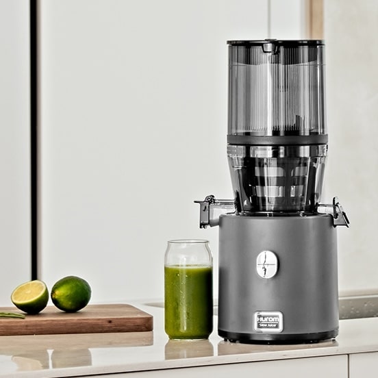 Hurom H330P Charcoal Entsafter in einer hellen Küche, daneben ein Glas grüner Saft und Limetten.