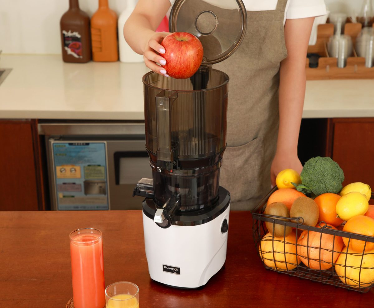 Eine Person legt einen ganzen Apfel in die Trommel, mit einem Glas Saft vor dem Entsafter und einem Korb mit Obst und Gemüse rechts.