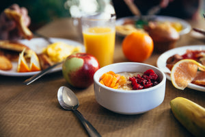 Eine komplett gedeckte Frühstückstafel mit Tellern voller gesunder Frühstücksoptionen, Schalen mit Müsli und frischem Obst. Auf dem Tisch liegen auch ganze Früchte wie Bananen, Orangen und Äpfel, und in der Mitte des Bildes steht ein Glas frisch gepresster Orangensaft.