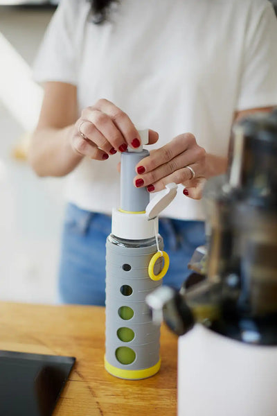 Frau mit roten Fingernägeln benutzt die Handpumpe nachdem sie die Flasche mit grünem Saft auf einer Holztheke gefüllt hat. Neben der Flasche steht ein Entsafter, aus dem der frische Saft stammt.