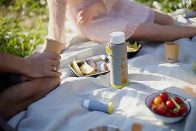 Mann und Frau sitzen auf einer Picknickdecke mit einer Pika Vakuumflasche zwischen ihnen, einer Schüssel mit frischen Tomaten und einem Tablett mit Ananasscheiben und anderen Früchten wie Orangen.